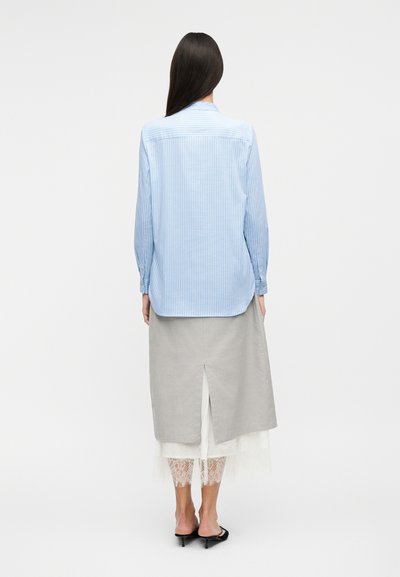 Γυναίκα με μακριά σκούρα μαλλιά φοράει γαλάζιο πουκάμισο με ρίγες, γκρι μίντι φούστα με άνοιγμα στο πίσω μέρος, λευκά δαντελένια παντελόνια και μαύρα σανδάλια με τακούνι, στέκεται με την πλάτη προς τα εμάς.