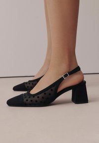 Musta slingback-kengät, joissa on paksu korko, verkkomateriaalia pilkullisella kuviolla, teräväkärkinen kärki ja hopeinen solki.