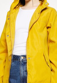 Žlutá kapucí bunda vyrobená z měkkého, texturovaného materiálu. Obsahuje přední zip, patentky a boční kapsy. Nositá přes bílý top.
