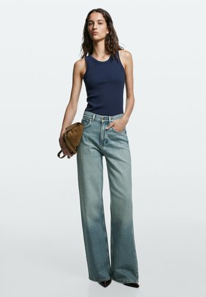 Kvinde med langt mørkt hår iført en navy ærmeløs top, brede lyseblå jeans, spidse mørke sko og holder en brun ruskindstaske.