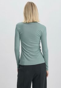 Maglia a maniche lunghe in morbido e elasticizzato tessuto verde teal, con una silhouette aderente e una texture liscia. Presenta un colletto rotondo e un design senza cuciture.