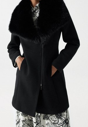 Manteau noir avec grand col en fourrure, fermeture éclair devant et poches latérales, porté sur une robe blanche à motif floral noir. Mains dans les poches.
