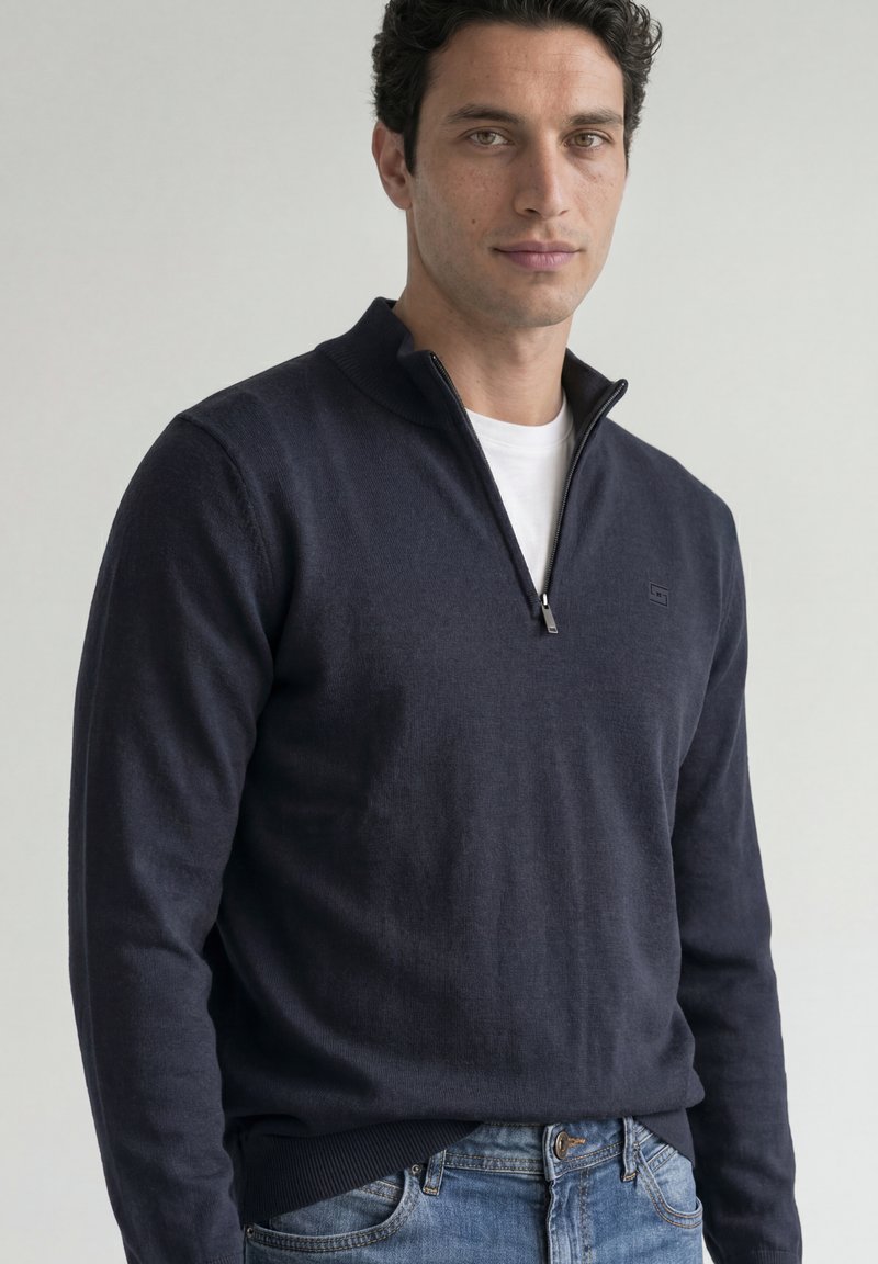 Homme aux cheveux courts, bouclés et foncés, portant un pull bleu marine à fermeture éclair quart sur une chemise blanche et un jean bleu, debout devant un fond uni.