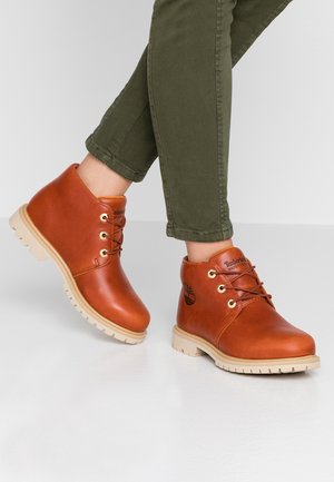 Person iført brune vandtætte læder Timberland-ankelstøvler med beige såler og grønne bukser, stående på et hvidt gulv.