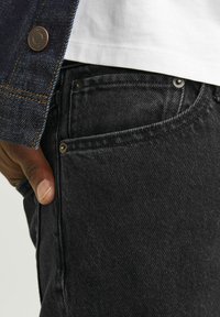 Vaqueros de mezclilla negra con una superficie texturizada, que presentan remaches metálicos, bolsillos laterales y una mano descansando sobre la zona del muslo.