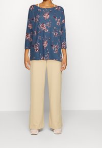 Sininen kukkakuosiset yläosa, jossa on kolme neljäsosaa hihat, yhdistetty beigeihin leveälahkeisiin housuihin. Kangas näyttää pehmeältä ja kevyeltä.