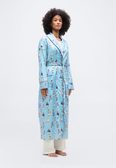 Γυναίκα με σγουρά μαλλιά φορώντας μακρύ μπλε ριγέ ρόμπα με σχέδια από κινούμενα σκυλάκια, δεμένη στη μέση, και άσπρο παντελόνι, όρθια χωρίς παπούτσια.