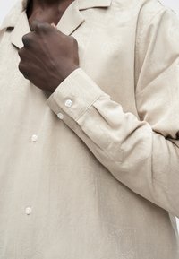 Beżowa koszula zapinana na guziki z delikatnym wzorem paisley, dopasowanym kołnierzykiem i długimi rękawami z mankietami na guziki. Gładka faktura tkaniny.