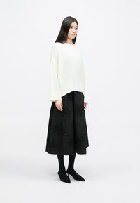 White knitted sweater with wide sleeves paired with a black floor-length skirt featuring floral patterns and beaded detailing. Black tights and flats.