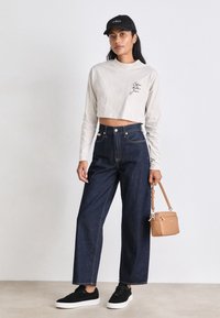 Chemise beige à manches longues raccourcie avec texte noir, jean large taille haute en denim foncé, baskets noires et sac à main beige. Casquette noire.