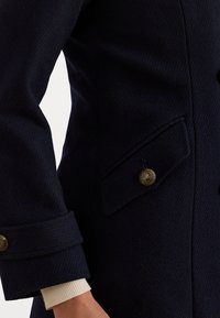 Abrigo de lana azul marino con una tela texturizada, cierres de botones dorados y un bolsillo lateral con solapas. Las muñecas están dobladas con un revestimiento de color crema.