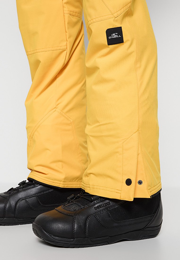 Gelbe Schneehosen mit verstärkten Nähten und einer Seitentasche; kombiniert mit schwarzen, strukturierten Low-Top-Schuhen, die ein Logo an der Seite tragen.