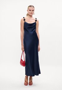 Rochie maxi din satin bleumarin cu decolteu cowl și accente florale pe bretele. Asortată cu o geantă roșie și sandale roșii cu barete.