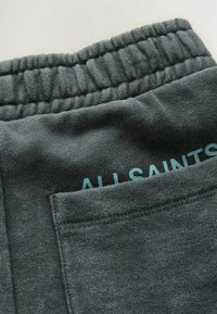 Tummanharmaat collegehousut, joissa onTexturoitu kangas, joustava vyötärö ja takatasku, jossa on teal-värillä painettu "ALLSAINTS".