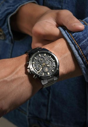 Orologio cronografo nero e argento al polso, con quadrante nero testurizzato, dettagli dorati e bracciale in acciaio inossidabile. Tessuto di denim visibile.