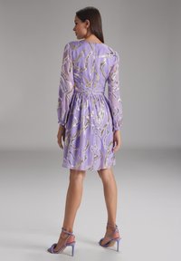Robe violette avec de longues manches transparentes, ornée de motifs dorés en spirale, taille cintrée et jupe évasée. Portée avec des talons violets assortis.