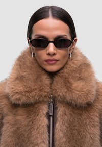 Cappotto di pelliccia marrone con un ampio colletto, dotato di chiusura a zip. Il modello indossa occhiali da sole rettangolari neri e orecchini a cerchio argentati.