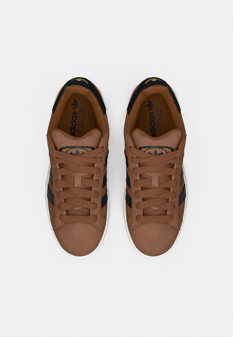 Zapatillas marrones con textura suave, con rayas negras y cordones, suela blanca y un detalle de logo dorado en la lengüeta.