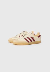 Cremefarvede og røde lædersneakers med tre røde striber, rund tå, tekstureret gummi-sål og logo på siden. Afslappet design.
