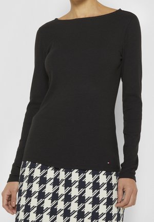 Femme portant un haut noir moulant à manches longues et une jupe à motif pied-de-poule noir et blanc, debout devant un fond uni.