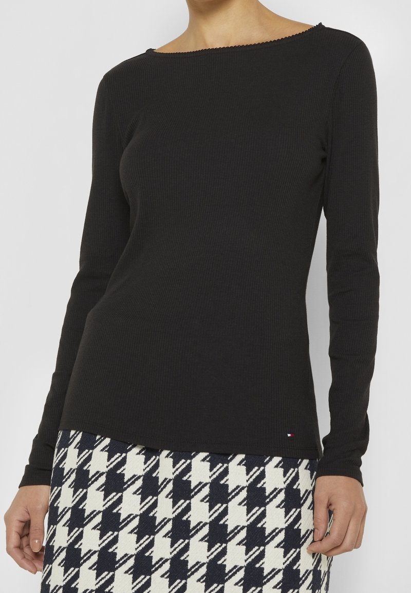 Femme portant un haut noir moulant à manches longues et une jupe à motif pied-de-poule noir et blanc, debout devant un fond uni.