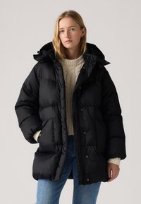 Black puffer jacket with a hood, featuring a quilted design, snap buttons, and side pockets, paired with a cream sweater and blue jeans.