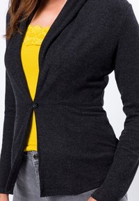 Cardigan noir à manches longues avec un col châle, une fermeture à un bouton et une taille cintrée. En dessous, un haut brillant jaune avec des lacets est visible.