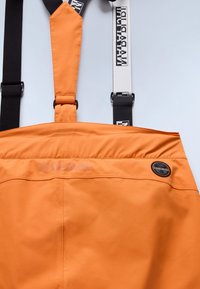 Oranssit vedenpitävät haalarit, joissa on mustat ja valkoiset säädettävät hihnat. Varustettu logopatchilla ja huomaamattomalla bränditekstillä, joka on kohotettu selkäpuolelle.