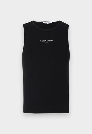 Jeune homme aux cheveux bouclés portant un haut noir sans manches, un pantalon noir ample et des baskets blanches, debout devant un fond clair uni.