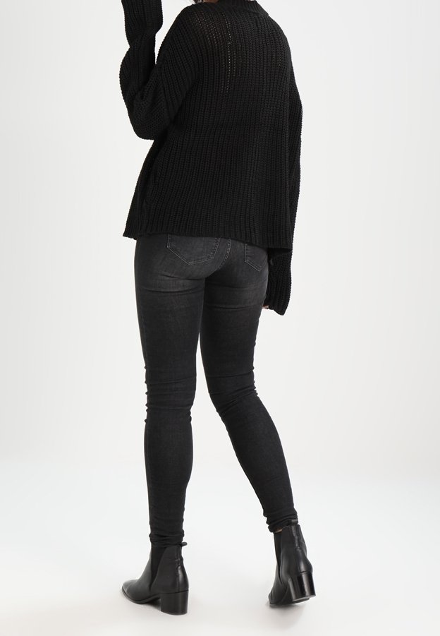 Personne portant un pull en grosse maille noire, un jean skinny noir et des bottines noires à talons, debout sur un fond blanc uni.