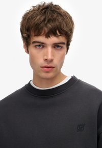 Sudadera negra con cuello redondo acanalado y logotipo bordado en el pecho, llevada sobre una camisa blanca, textura suave.
