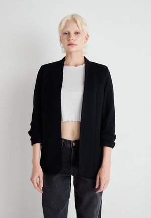 Černý blazer s přiléhavým střihem, s klopovým límcem, dlouhými rukávy a otevřeným předním dílem, nošený přes bílý žebrovaný crop top.