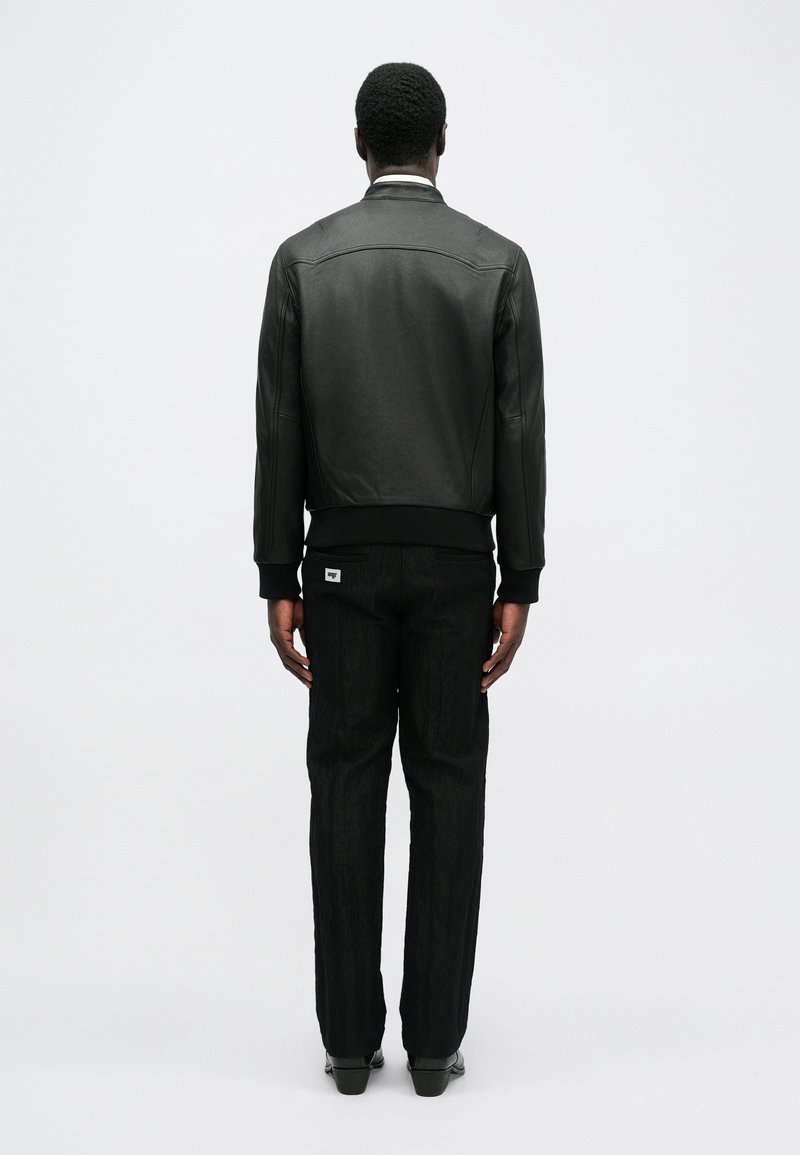 Black leather bomber jacket with a ribbed hem, featuring smooth texture and minimalist design. Paired with textured black trousers.