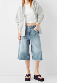 Felpa grigia chiara con zip sopra una canotta bianca, abbinata a pantaloni culotte in denim chiaro a gamba larga e sandali neri con suola piatta.