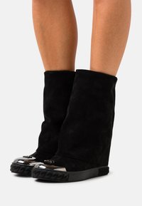 Botines de ante negro con diseño plegado, punta redondeada y detalles metálicos. Presentan una textura trenzada en la suela.