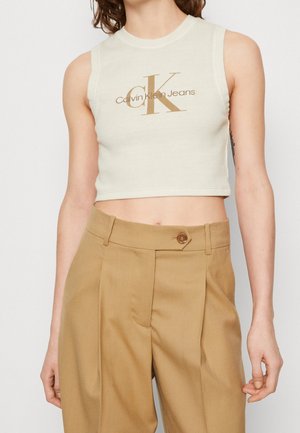 Osoba nosząca bezrękawnikowy biały krótki top z logo "Calvin Klein Jeans" oraz wysokie beżowe plisowane spodnie zapinane na guzik.
