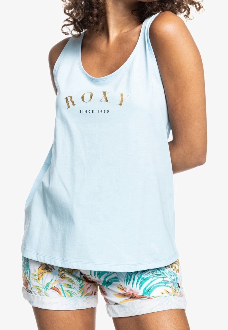 Femme portant un débardeur bleu clair avec le texte "ROXY Since 1990" et un short blanc avec des motifs de feuilles tropicales colorées.