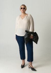 Beige blouse met V-hals en koord, marineblauwe cropped broek met split onderaan, zwarte leren clutch tas en zwarte puntige hakken.