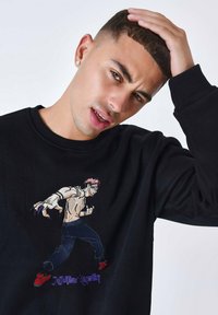 Sweat-shirt noir avec un graphique brodé d'un personnage aux cheveux roses et éléments bleus, accompagné de chaussures rouges.