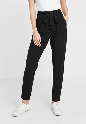 Vrouw die zwarte, hooggetailleerde, taps toelopende broek draagt met een paperbag-tail en een gestrikte ceintuur, gecombineerd met witte sneakers en een wit mouwloos topje.