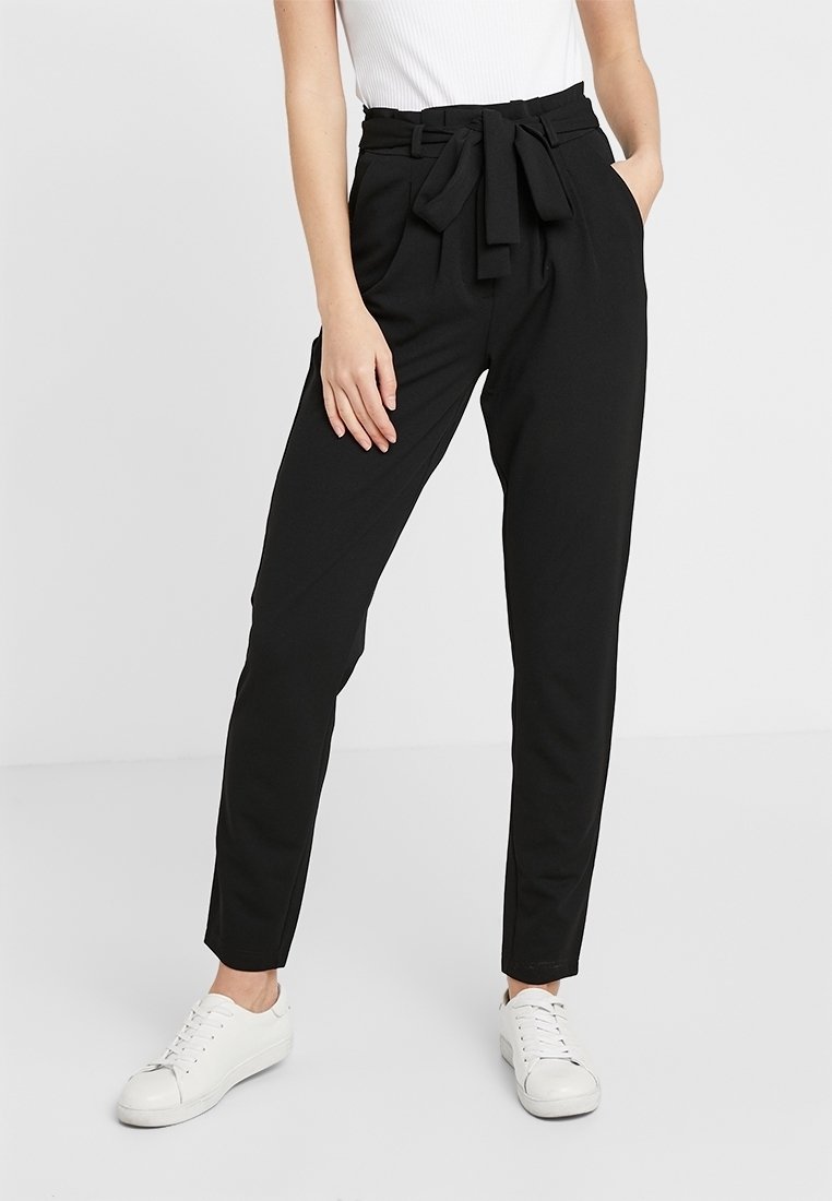 Femme portant un pantalon noir taille haute cintré avec une taille plissée et une ceinture nouée, associé à des baskets blanches et un haut blanc sans manches.