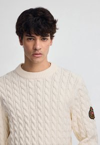 Cremefarbener Kabelstrickpullover mit geripptem Kragen und Bündchen. Verfügt über einen runden Ausschnitt und ein Emblem an der linken Schulter. Strukturiertes Design im gesamten Pullover.