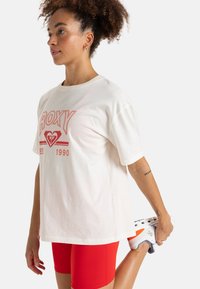 T-shirt beige clair avec le logo rouge "ROXY" et le texte "EST 1990". Coupe décontractée, col rond, associé à un short rouge et des chaussures de sport multicolores.