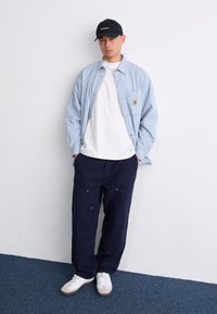 Lysblå stribet skjorte med knapper over en hvid t-shirt, parret med navy bukser og hvide sneakers med beige detaljer; sort kasket inkluderet.