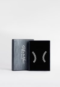 Silvergeometriska örhängen med diamantformade utskärningar, som visas i en strukturerad mörkblå låda med svart sammet på insidan.