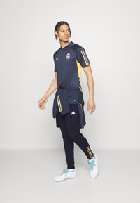 Jersey deportivo azul marino con rayas amarillas y púrpuras, combinado con pantalones joggers azul marino y zapatillas blancas con cordones azul celeste.
