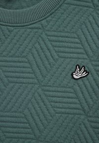 Tmavozelený sveter s textúrovaným šestuholníkovým vzorom, rebrovaným výstrihom a vyšitou emblémou vtáka v bielej a čiernej farbe.