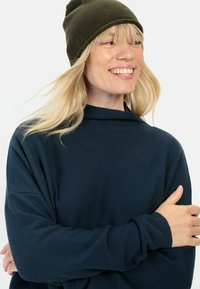 Marineblauwe trui met hoge kraag en ribbelstructuur, gecombineerd met een donkergroene gebreide beanie. Het model heeft lang, recht blond haar.