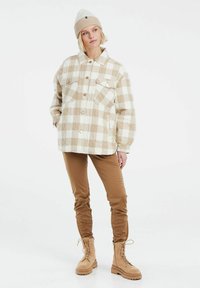 Ruudullinen beige ja valkoinen flanellipaita-takki, jossa on kaksi etutaskua, yhdistettynä ruskeisiin housuihin ja vaaleisiin nauhakorkokenkiin, sekä beige neulottu pipo.