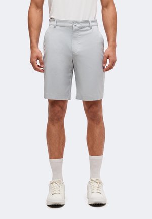 Homme portant un short gris clair mi-genou, des baskets blanches, des chaussettes blanches et une chemise blanche, debout devant un fond uni.